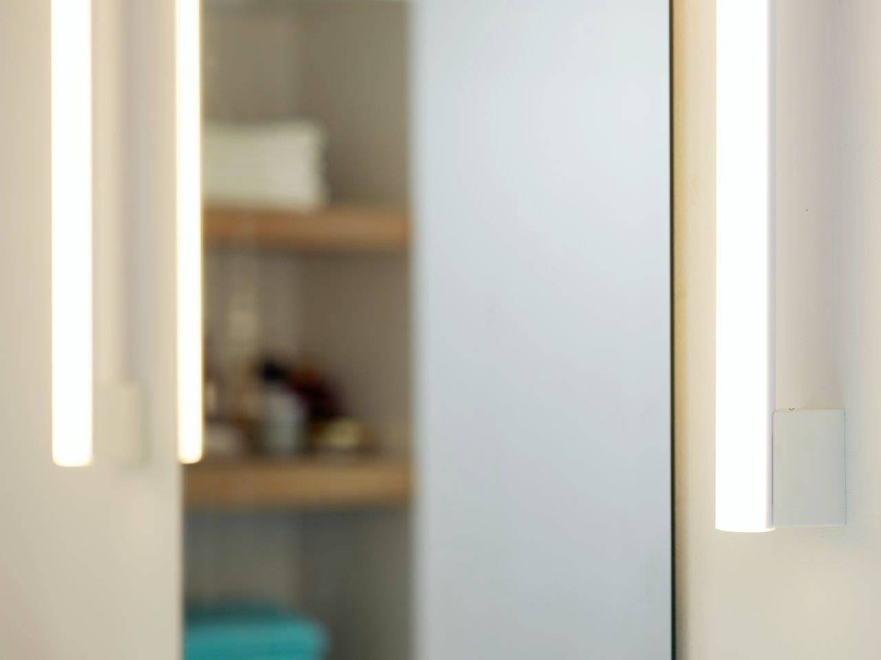 Osvetlené zrkadlo s dvomi vertikálnymi svetlami po stranách. V pozadí sú rozmazané police s rôznymi predmetmi.