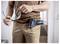 Osoba v pracovnom oblečení drží bezpečnostné okuliare, pričom bezdrôtová vŕtačka je vložená do vrecka vpredu.