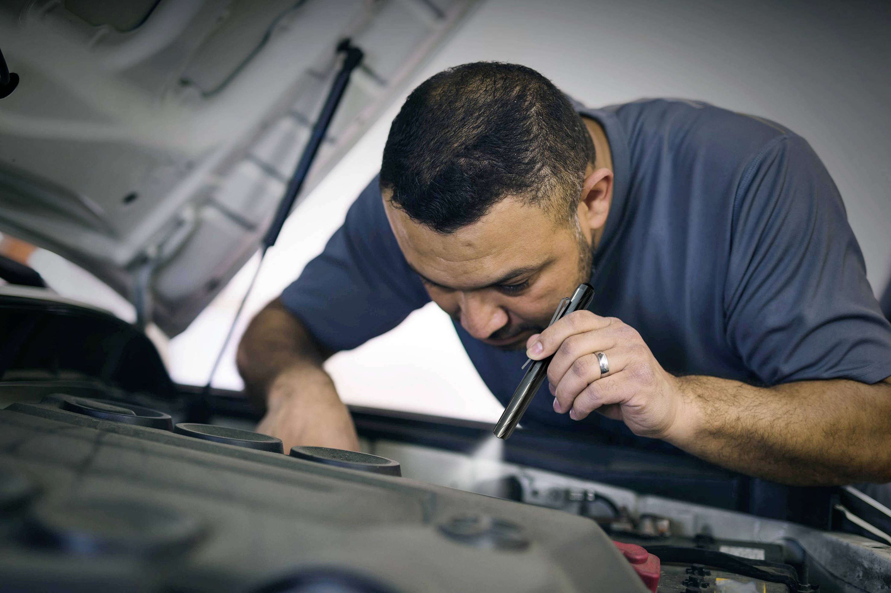 Mechanik skúma automobil pod otvorenou kapotou. Používa malú baterku na kontrolu motorového priestoru.