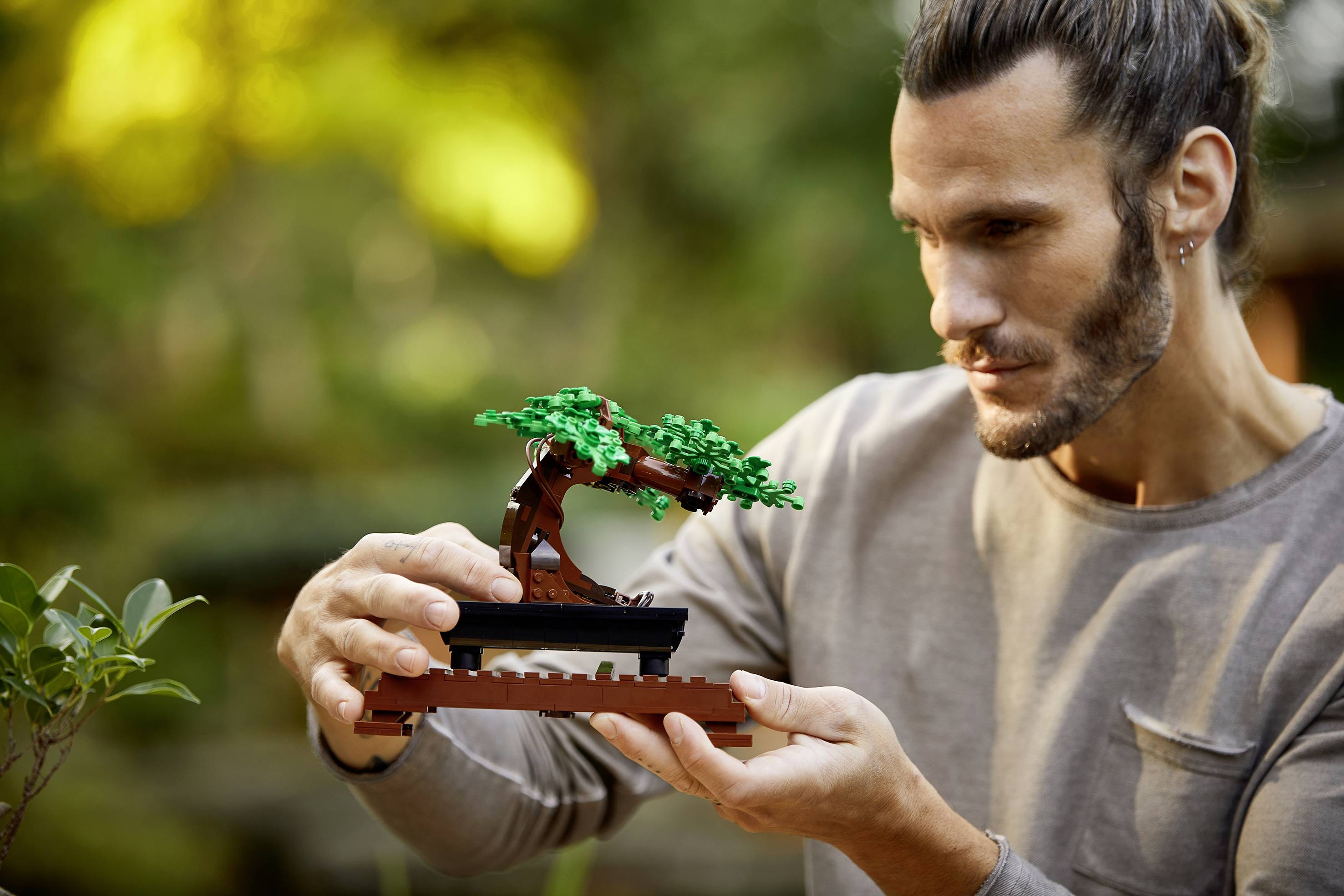 Muž drží malý Lego bonsajový model stromu vonku. V pozadí sú rozmazané listy.