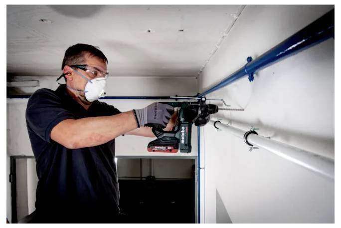 Osoba, ktorá má na sebe ochranné okuliare a masku, používa elektrickú vŕtačku na vyvŕtanie otvoru do steny, pričom sa sústreďuje na svoju úlohu v malej miestnosti.