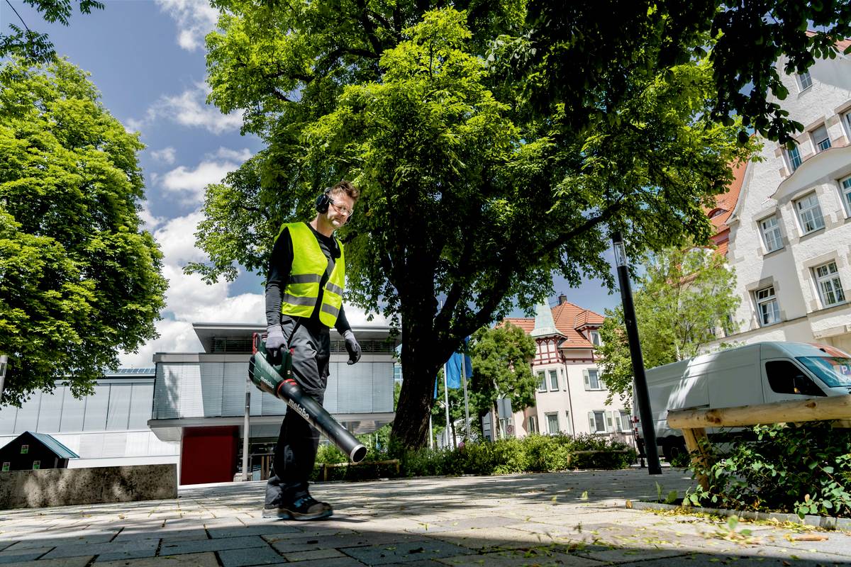 Muž v pracovnom oblečení používa fukár na lístie na chodníku. V pozadí sú viditeľné stromy a budovy.