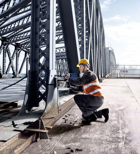 Robotník v ochrannej prilbe a reflexnej veste pracuje na nosníku mosta vŕtačkou počas slnečného počasia.