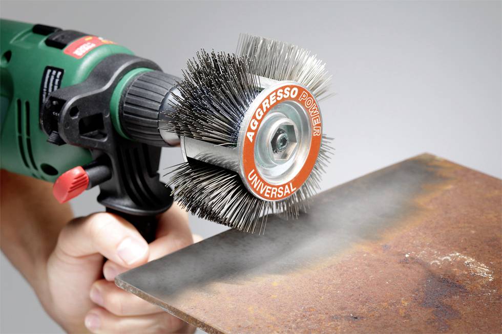Jedna ruka drží elektrickú drôtenú kefu s nápisom 'Aggresso Power Universal', ktorá odstraňuje hrdzu z kovovej časti.