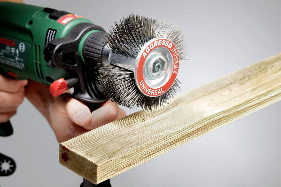 Osoba brúsi kus dreva elektrickým brúsnym kefovým nástavcom. Brúsna kefa má nápis 'Aggresso Power Universal'.