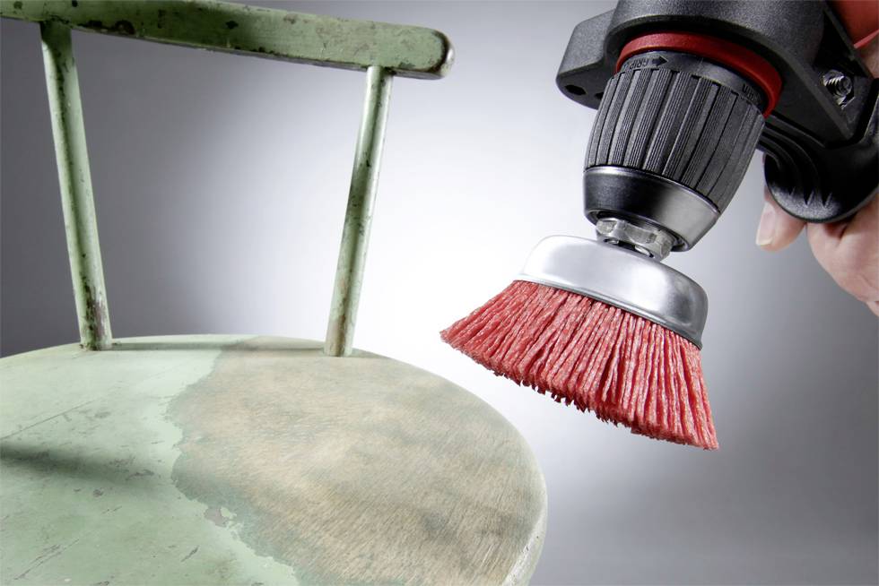 Osoba brúsi sedadlo starého kovového stoličky červenou rotačnou kefou, aby ho renovovovala.