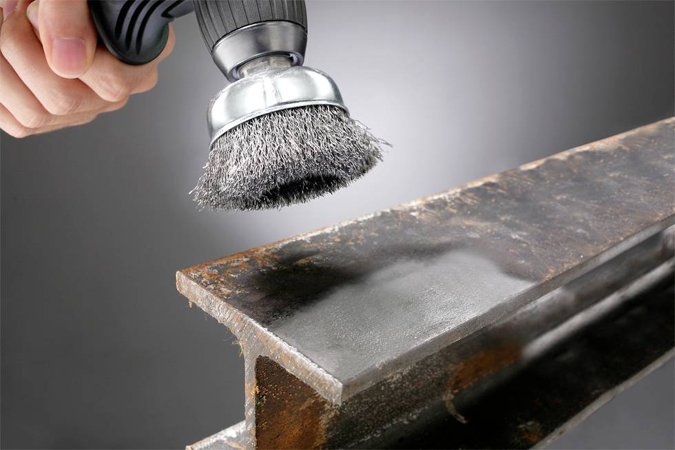 Osoba používa drôtenú kefu s kovovými štetinami na odstránenie hrdze z kovového nosníka a vyhladenie povrchu.