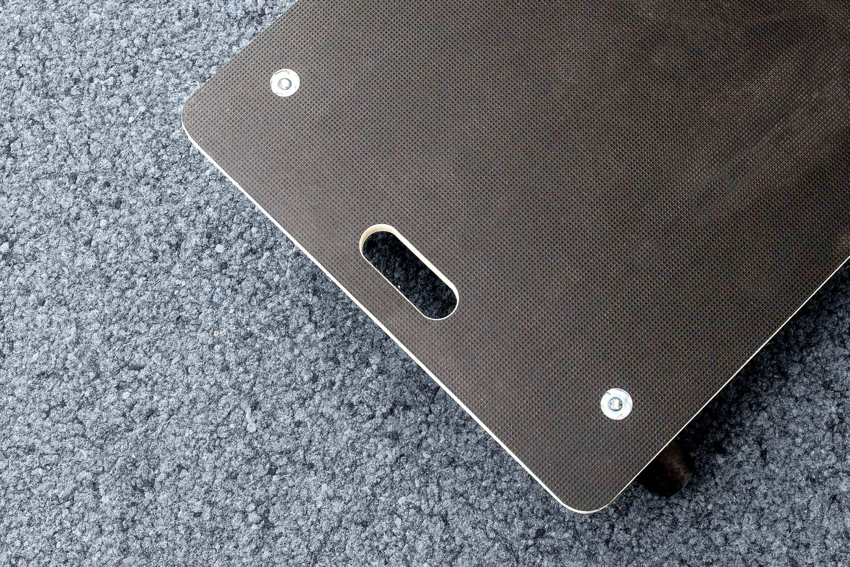 Obdĺžnikový skateboard deck leží na asfaltovom povrchu. Deck má otvor na uchopenie a dve viditeľné skrutky.