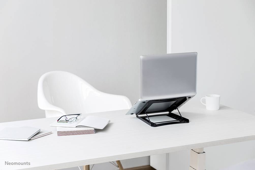 Laptop na výškovo nastaviteľnom stojane stojí na písacom stole. Vedľa ležia okuliare, knihy a šálka.