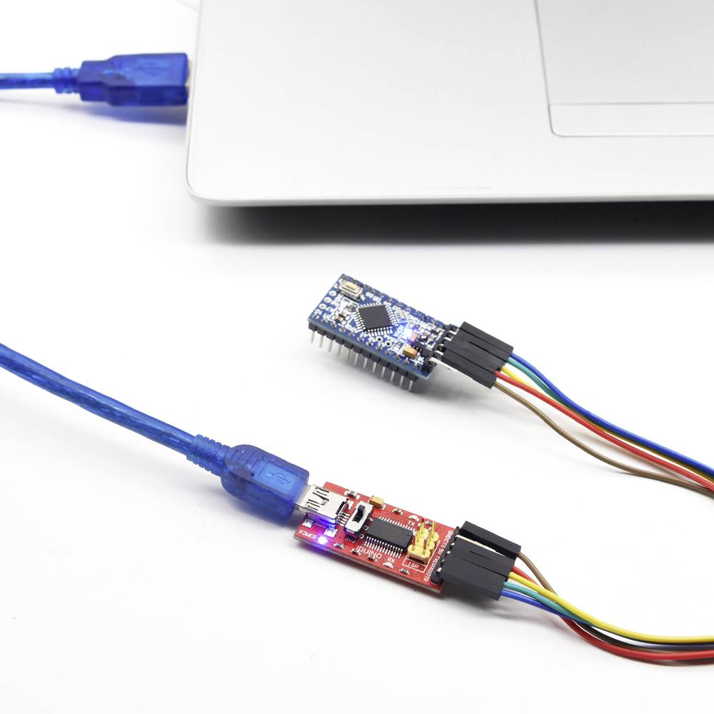 USB kábel, mikrocontroller a adaptér prepojené s laptopom. Scéna ukazuje setup pre elektronické programovanie alebo vývoj.