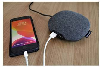 Smartfón sa nabíja cez USB kábel, pripojený k okrúhlej sivej nabíjačke na drevenej doske. Na obrazovke je zobrazený čas.