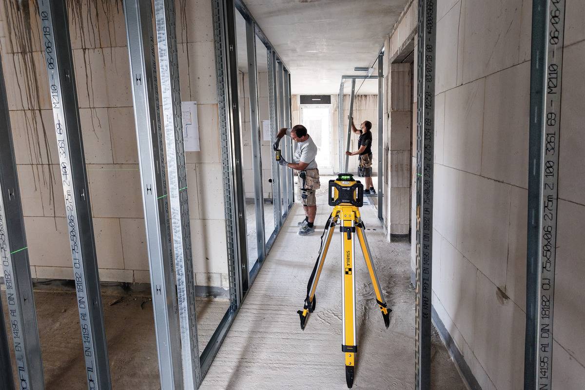 Dvaja stavebníci montujú kovové rámy v nedokončenej budove. V popredí stojí žlté laserové meracie zariadenie určené na presné vyrovnanie.