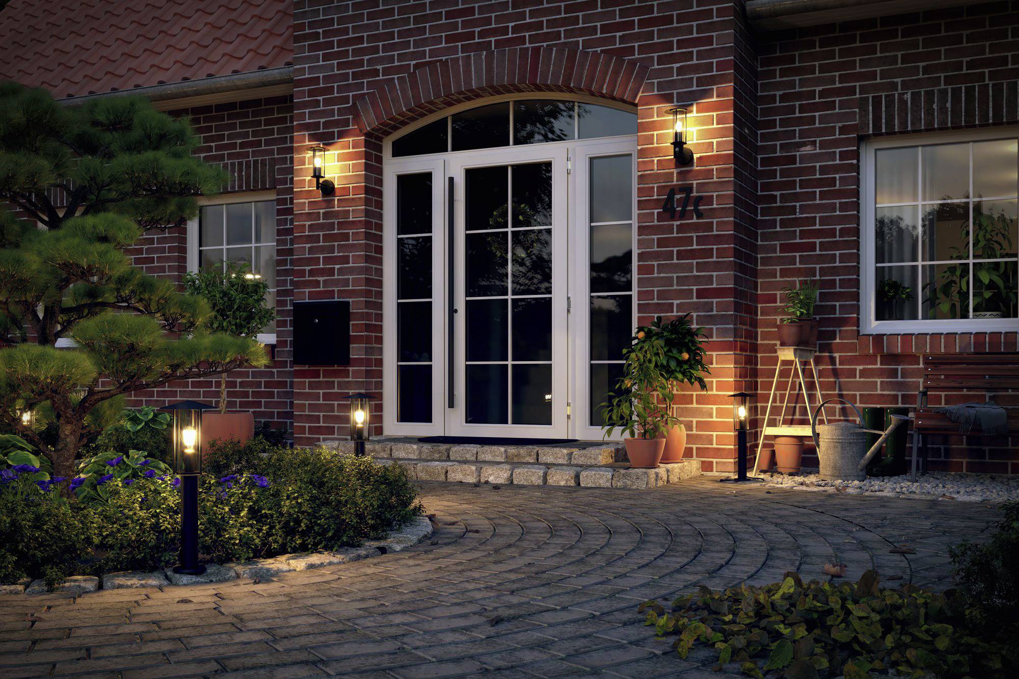 Vstup do tehlového domu za súmraku, osvetlený stenový svetlami. Udržiavaný chodník vedie cez malú záhradu.