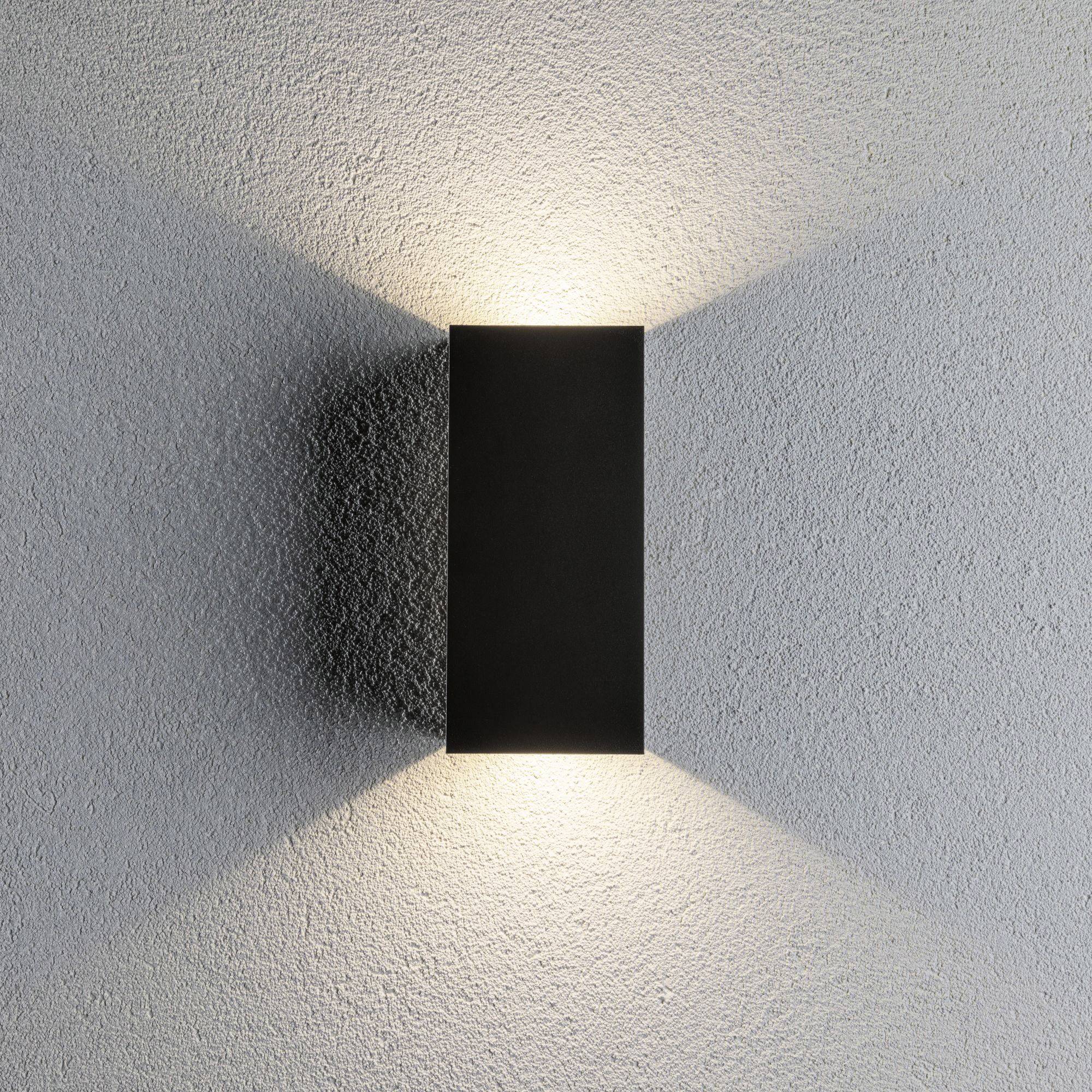 Obdĺžniková, čierna nástenná lampa vyžaruje svetlo nahor a nadol na sivej stene a vytvára atmosférické svetelné efekty.