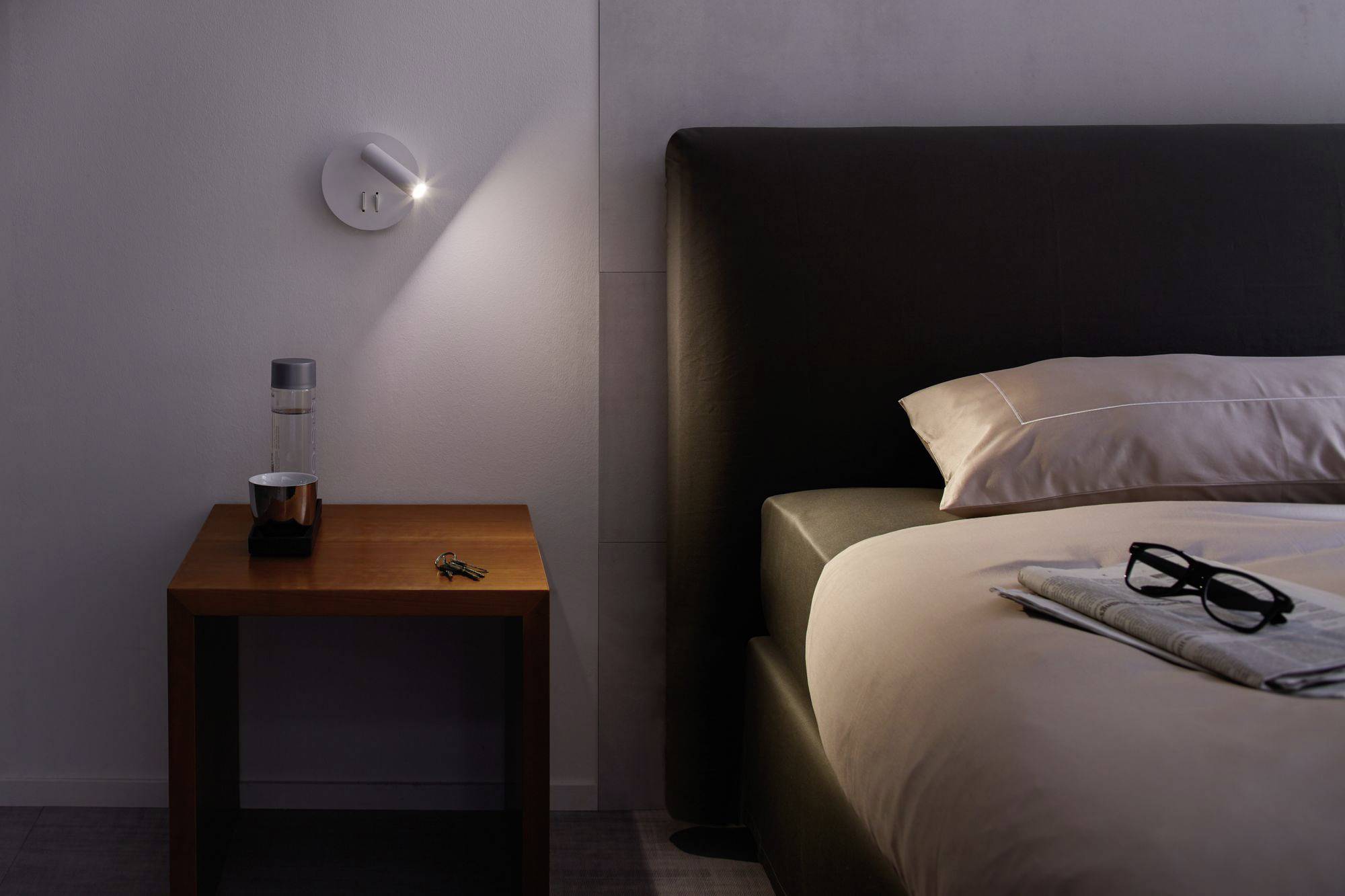 Nočný dojem hotelového priestoru s osvetlenou posteľou, nočným stolíkom s džbánom vody a pohárom. Kľúče ležia na stole.