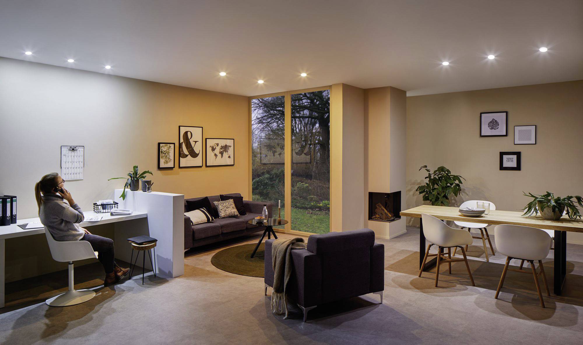 Moderne zariadená obývačka so stolom, pohovkou, jedálenským stolom a veľkými oknami, ktoré umožňujú výhľad do záhrady. Jedna osoba telefonuje pri stole.