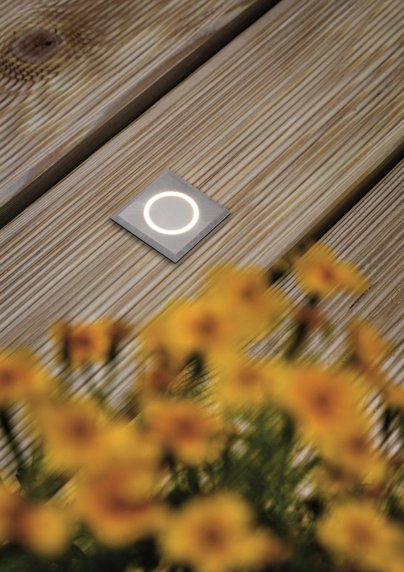 Svietiace štvorcové LED zapustené svetlo na drevenej terase, s rozmazanými žltými kvetmi v popredí.
