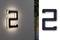 Osvetlené číslo domu '2' na stene, vedľa neho rovnaké číslo ako tmavá tabuľka na bielom pozadí.