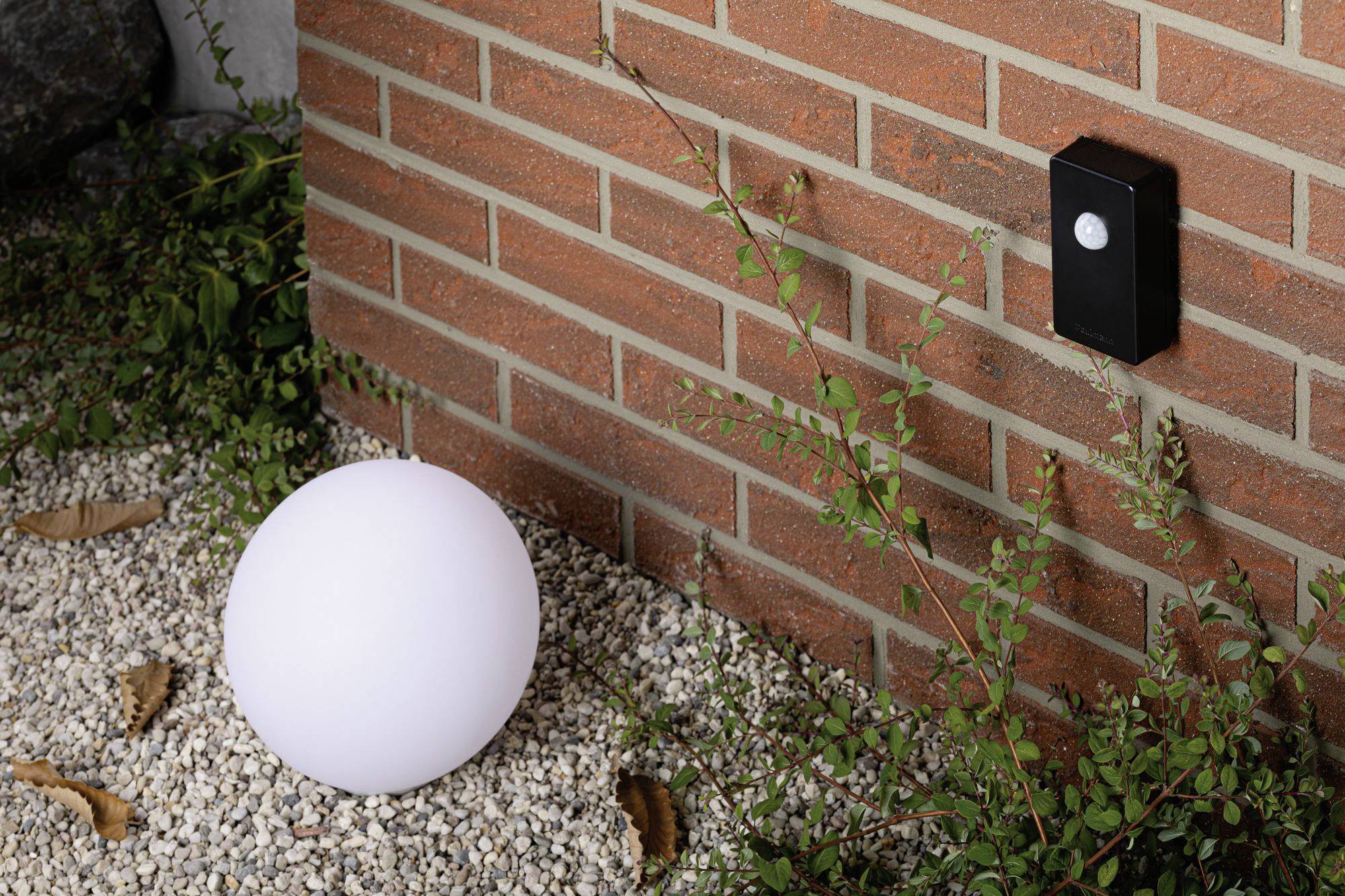 Biela, guľovitá lampa leží na štrkovej zemi vedľa tehlovej steny s čiernym vypínačom. Medzi nimi rastú zelené rastliny.