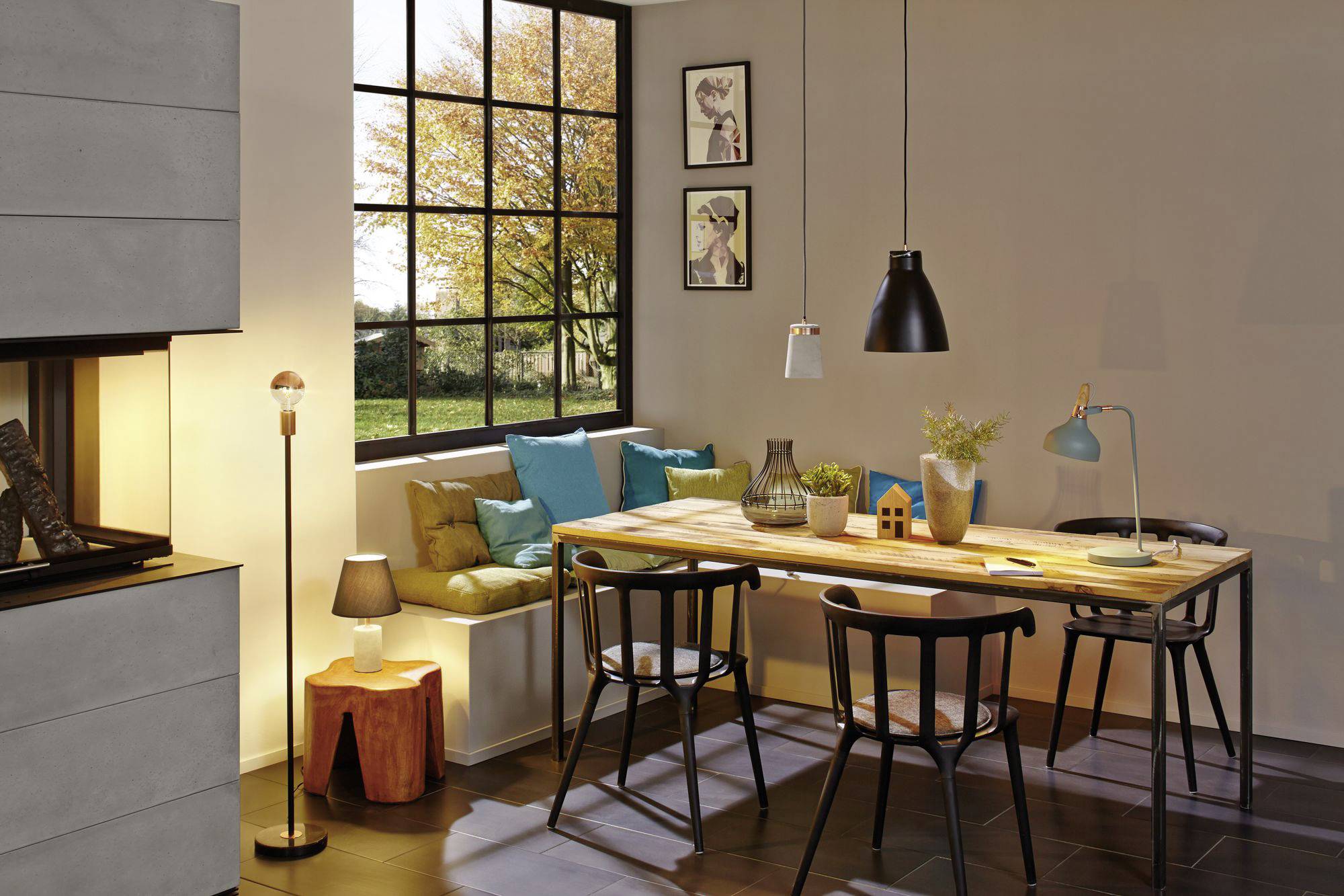 Jedáleň s veľkým dreveným stolom, štyrmi čiernymi stoličkami a farebnými vankúšmi na parapete. Veľké okná púšťajú dovnútra svetlo.