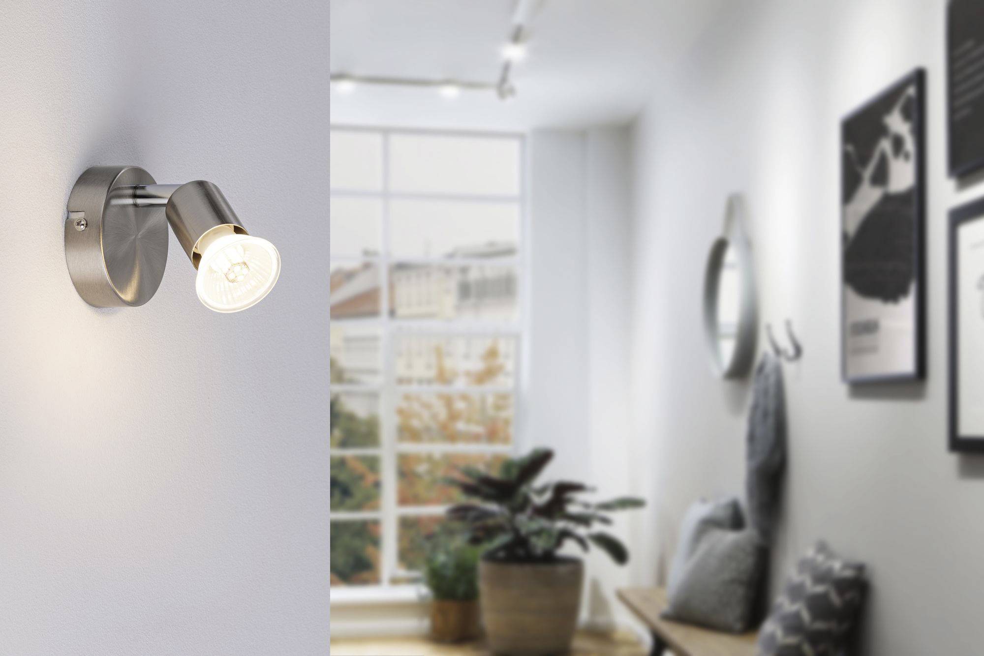 Nástenná lampa v modernej obývačke so svetlým denným svetlom, rastlinami a zarámovanými obrazmi na stene.