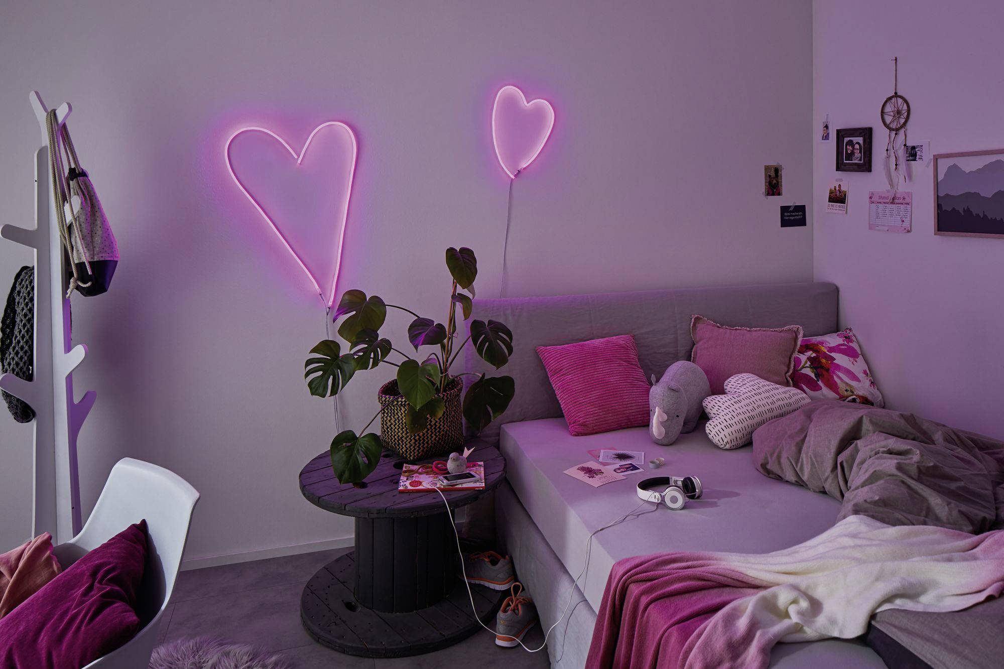 Útulná izba so sivou sedačkou, ružovými vankúšmi, rastlinami, neónovým svetlom v tvare srdca a dekoráciami na stene. Laptop a slúchadlá ležia na sedačke.