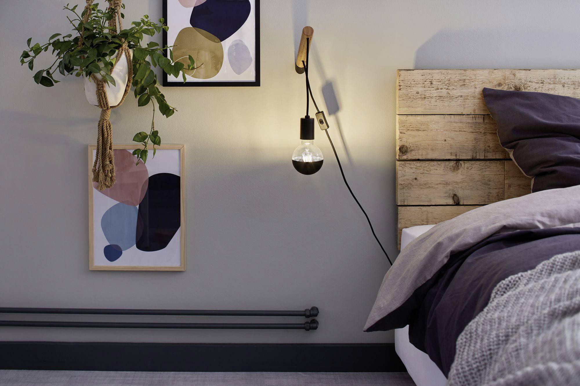 Útulná spálňa s drevenou posteľou, osvetlená lampou na stene. Izbu zdobia dva abstraktné umelecké diela a jedna rastlina.