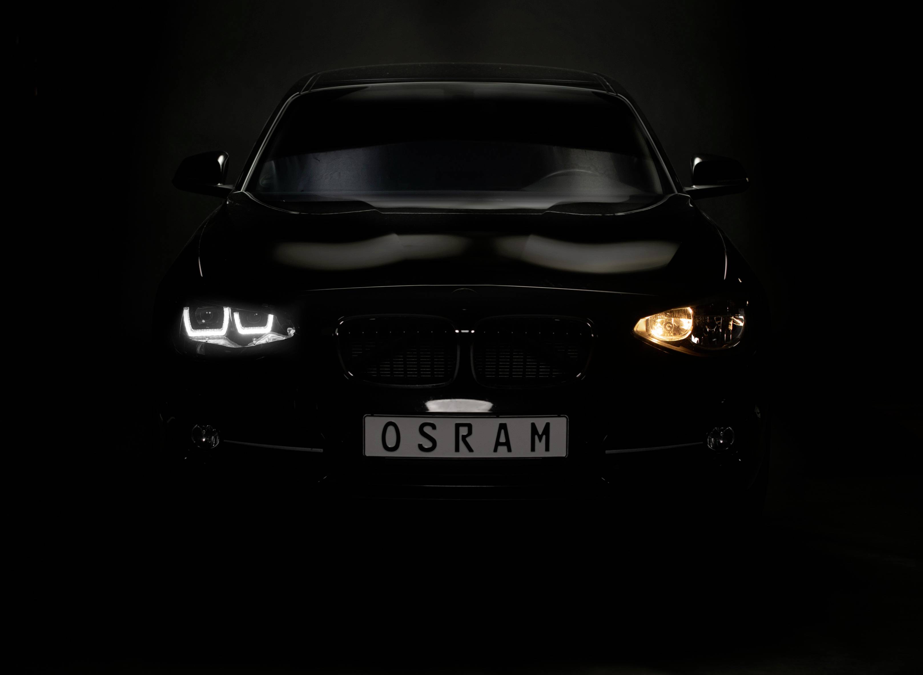 Čierne auto so zapnutým ľavým LED svetlometom a pravým žltým pozičným svetlom pred tmavým pozadím.