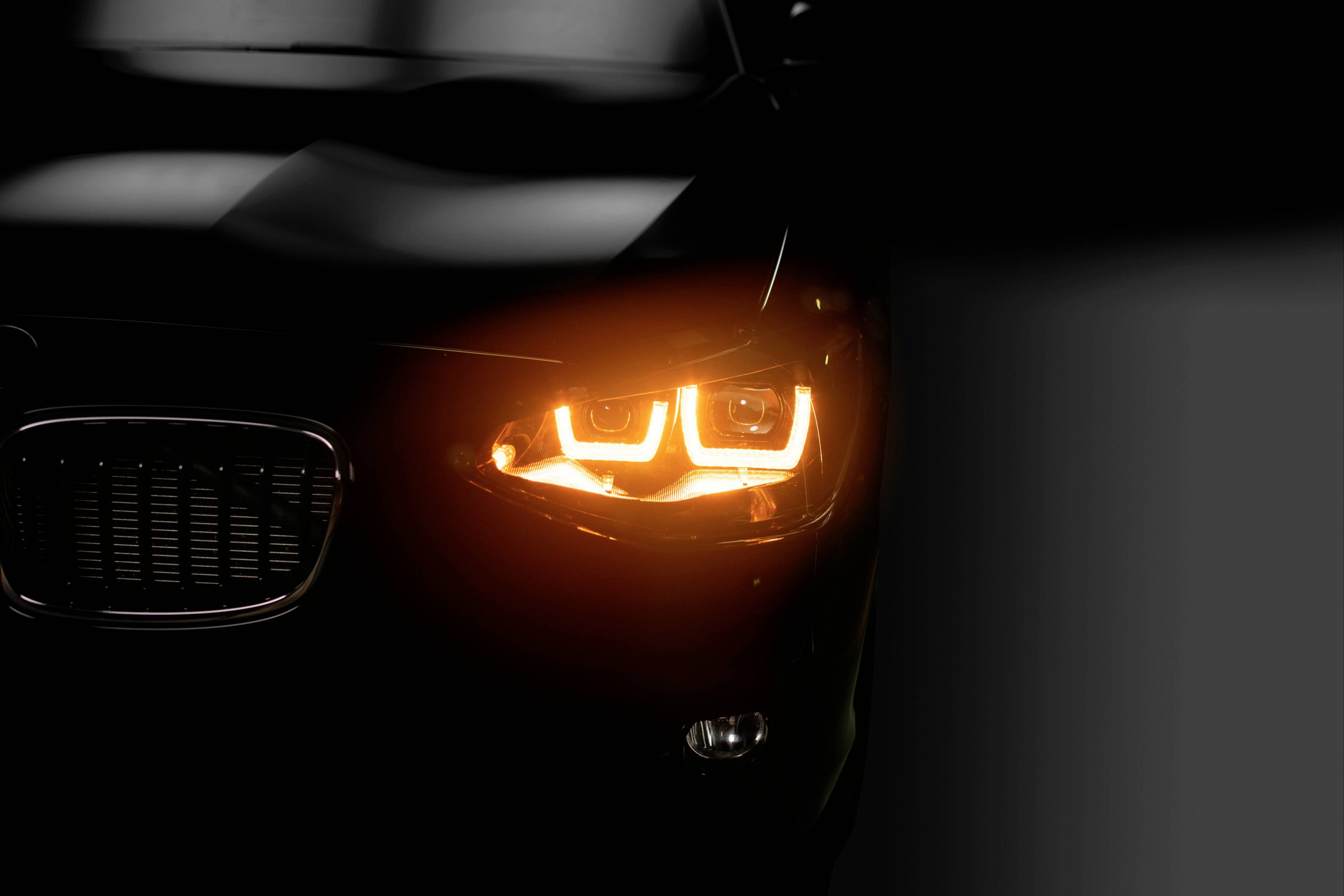 Detailný záber čierneho automobilu so svietiacim svetlometom na tmavom pozadí, prevažne viditeľná ľavá predná časť vozidla.