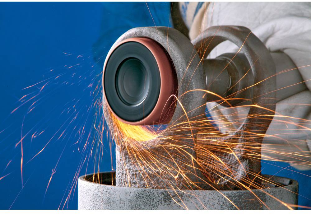 Z brúsneho kotúča sršia iskry, ktorý opracúva kov, nesený osobou v pracovných rukaviciach.