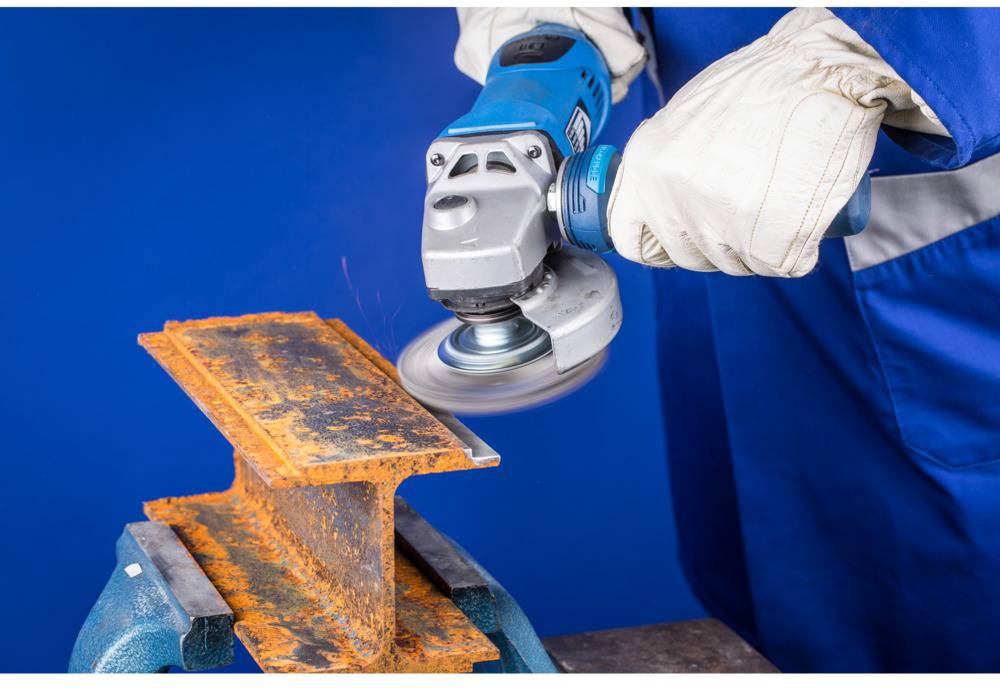 Osoba používa uhlovú brúsku na odstránenie hrdze z oranžového kovového kusu, ktorý je upevnený vo zveráku.