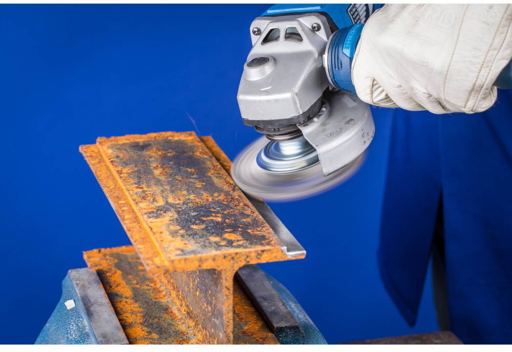 Osoba brúsi hrdzavú oceľovú dosku uhlovou brúskou. Doska je upevnená vo zveráku.