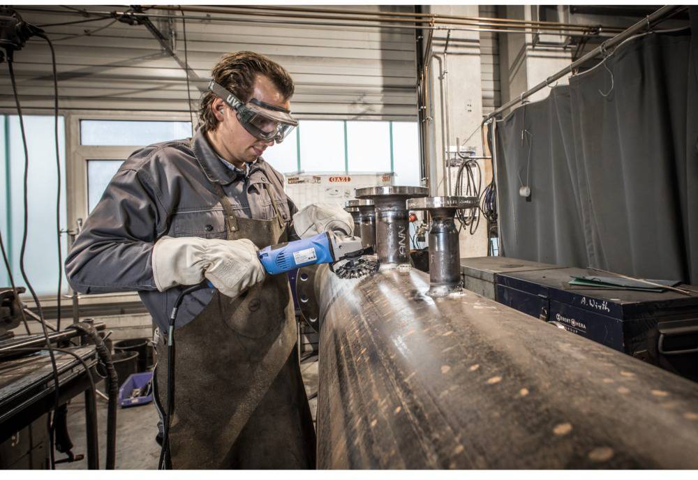 Muž v ochrannom oblečení a ochranných okuliaroch zvára veľký kovový valec v dielni.