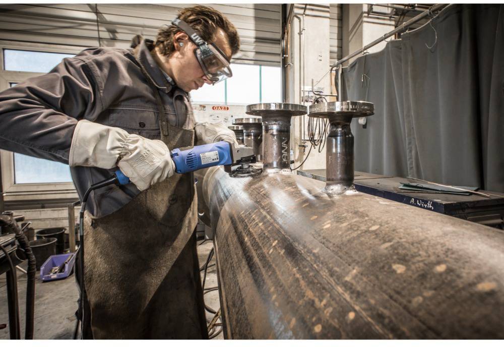 Remeselník v ochranných okuliaroch a rukaviciach brúsi veľkú kovovú rúru v dielni. V pozadí sú viditeľné rôzne nástroje a zariadenia.