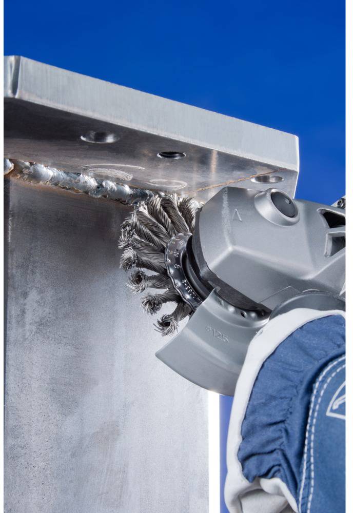 Osoba v ochrannom oblečení brúsi kovovitý zvárací šev brúskou pred čistým modrým pozadím.