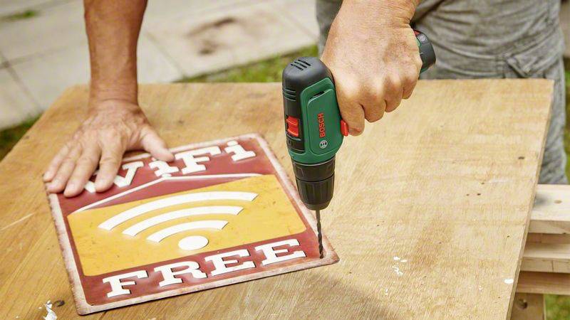 Osoba pripevňuje tabuľku s nápisom 'WiFi Free' akumulátorovým skrutkovačom na drevený stôl.