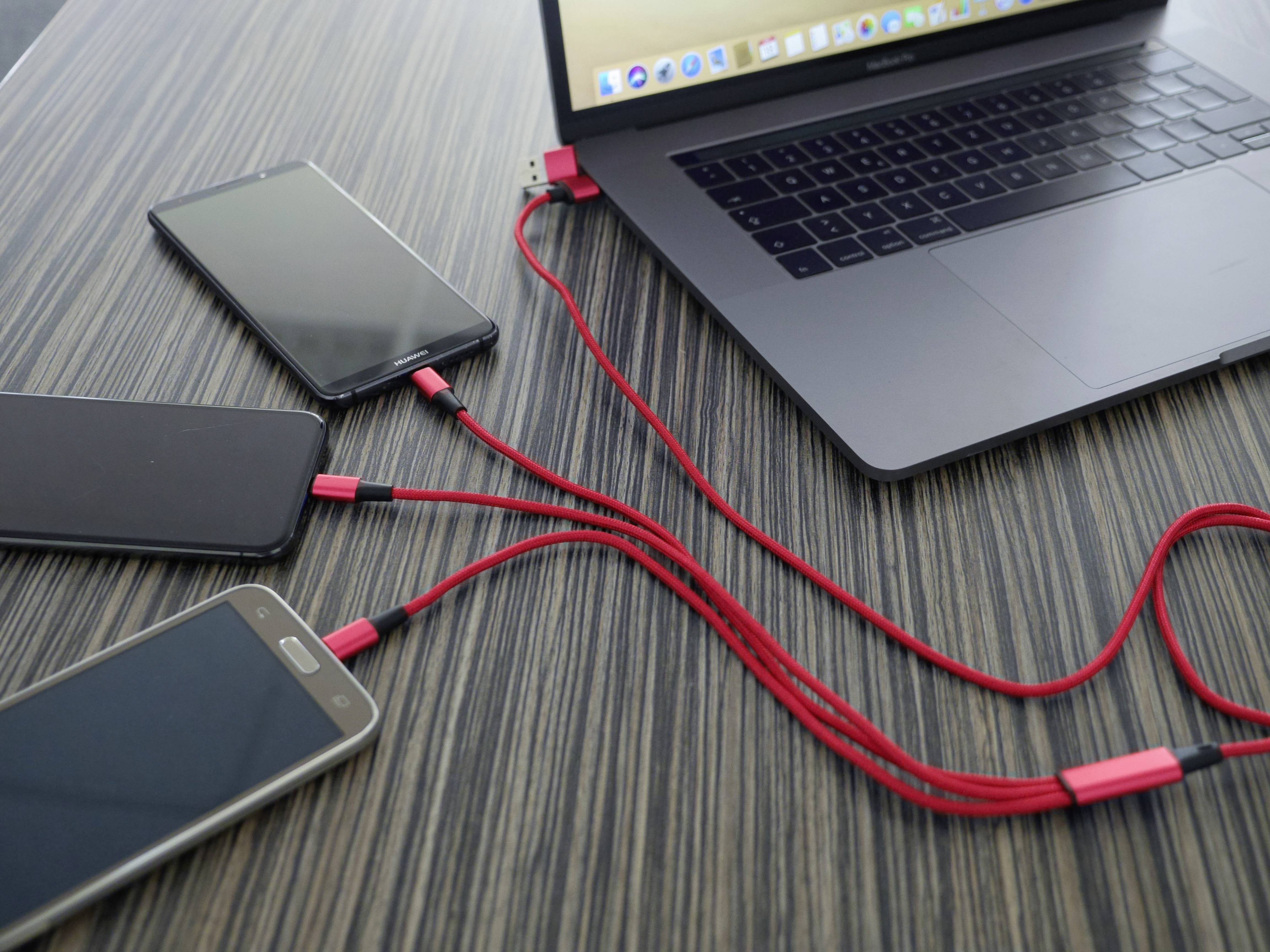 Laptop nabíja tri smartfóny červenými USB káblami na tmavom drevenom stole.