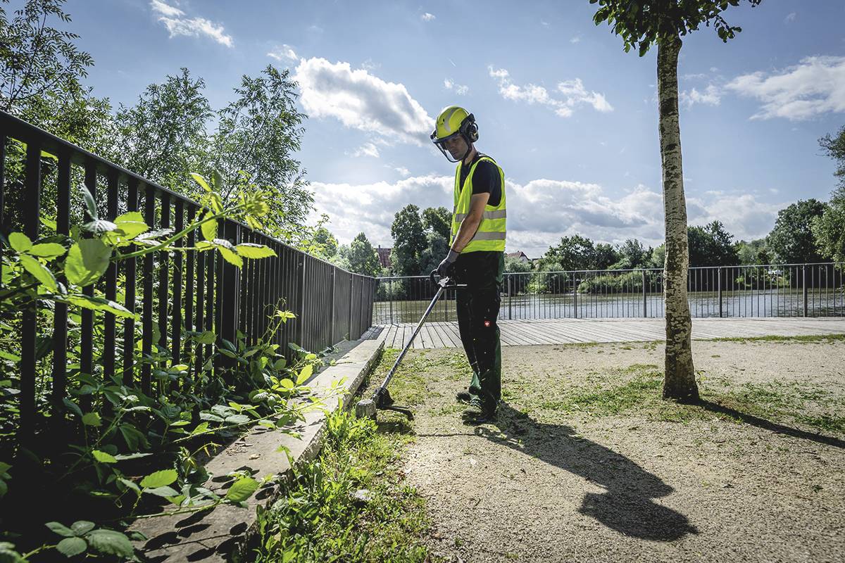 Osoba v bezpečnostnom oblečení používa vyžínač na strihanie rastlín pri plote v parku blízko rieky.