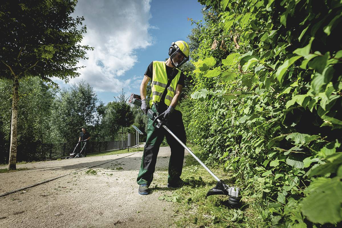 Robotník v ochrannom oblečení strihá kríky trimrom vedľa chodníka. V pozadí je ďalší robotník.