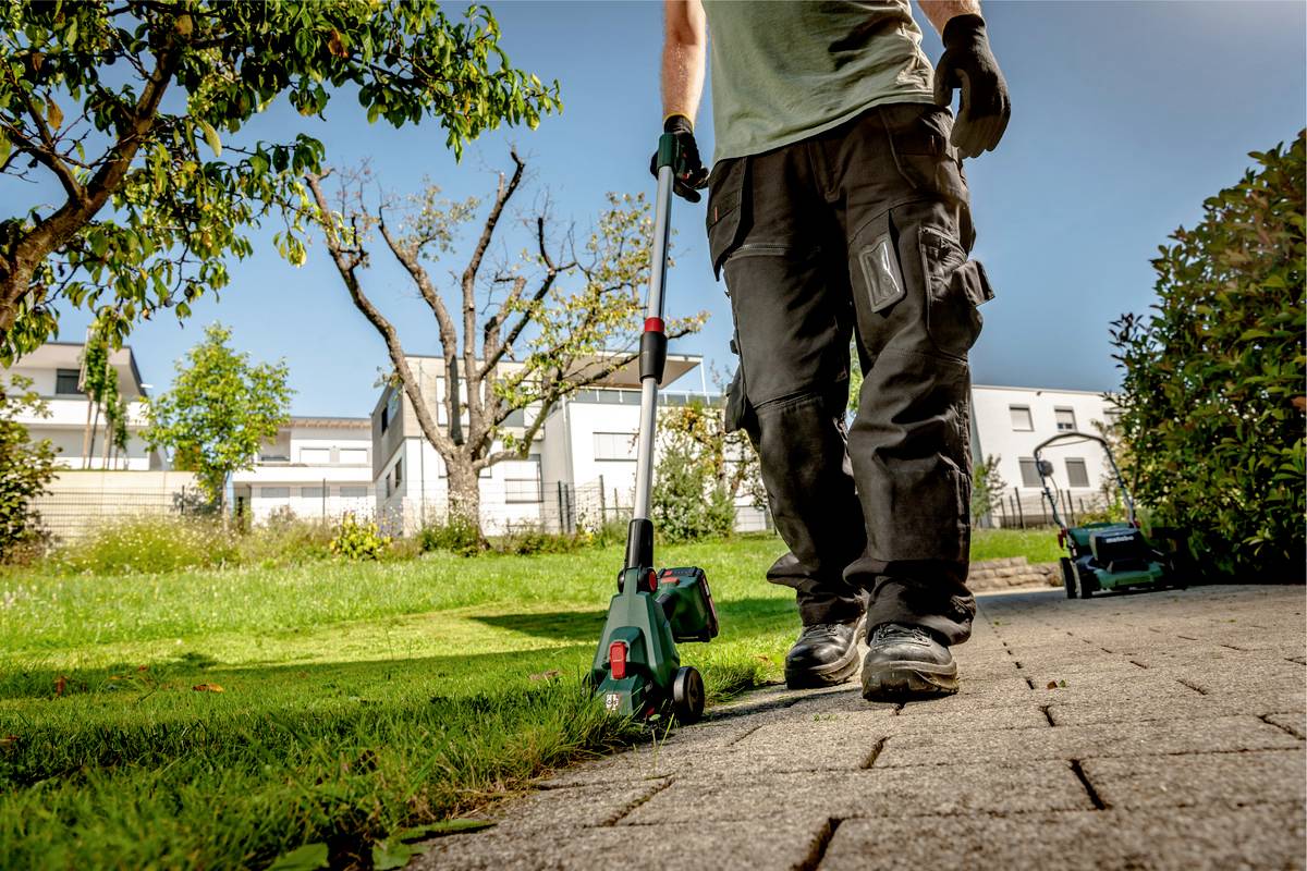 Človek obsluhuje elektrický vyžínač okrajov chodníka v slnečnom záhradnom prostredí pred modernými domami.