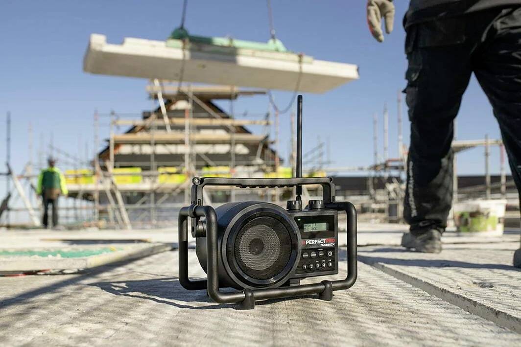 Stavenisko s prenosným rádiom v popredí, robotníci a lešenie v pozadí; budova vo výstavbe; slnečný deň.