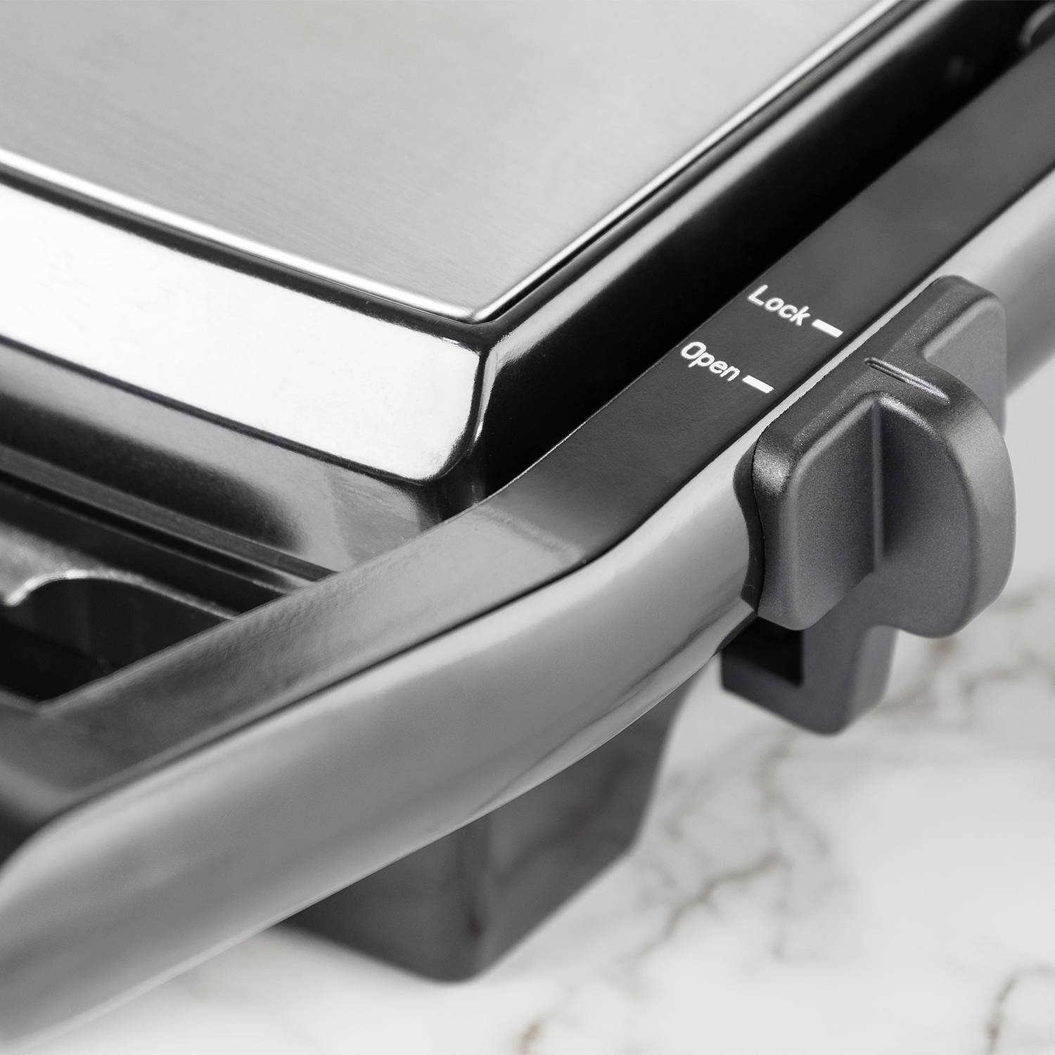 Detailný záber kuchynského prístroja s čiernou rukoväťou a tlačidlom, na ktorom je napísané 'Open' a 'Lock', umiestnený na bielom mramorovom povrchu.