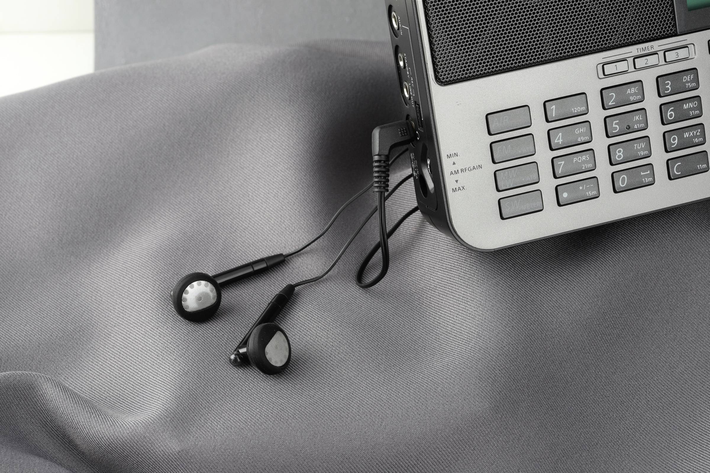 Strieborné rádio so slúchadlami na sivej látke. Rádio má tlačidlá, ovládač hlasitosti a zásuvku pre slúchadlá.