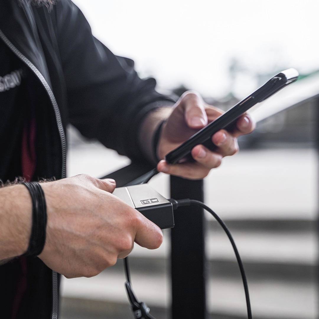 Osoba nabíja smartfón powerbankou počas pobytu vonku. Nie sú viditeľné žiadne ďalšie detaily ani kontext.