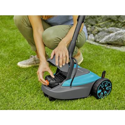 Osoba mení batériu bezdrôtového robotického kosačky na zelenej tráve, pričom robotický prístroj má modré akcenty.