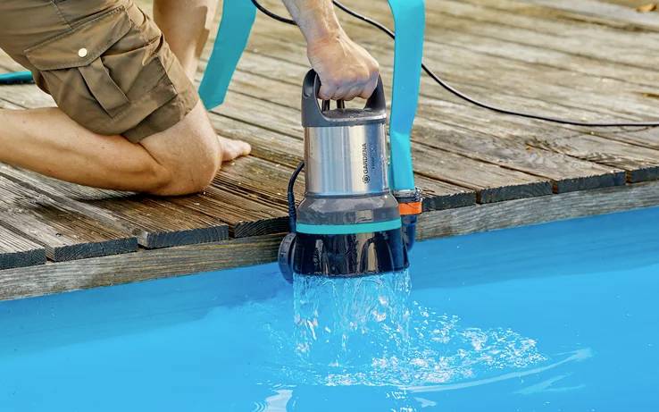 Osoba čerpá vodu z modrého bazéna pomocou elektrickej pumpy. Pumpa má telo z kovu a plastu.