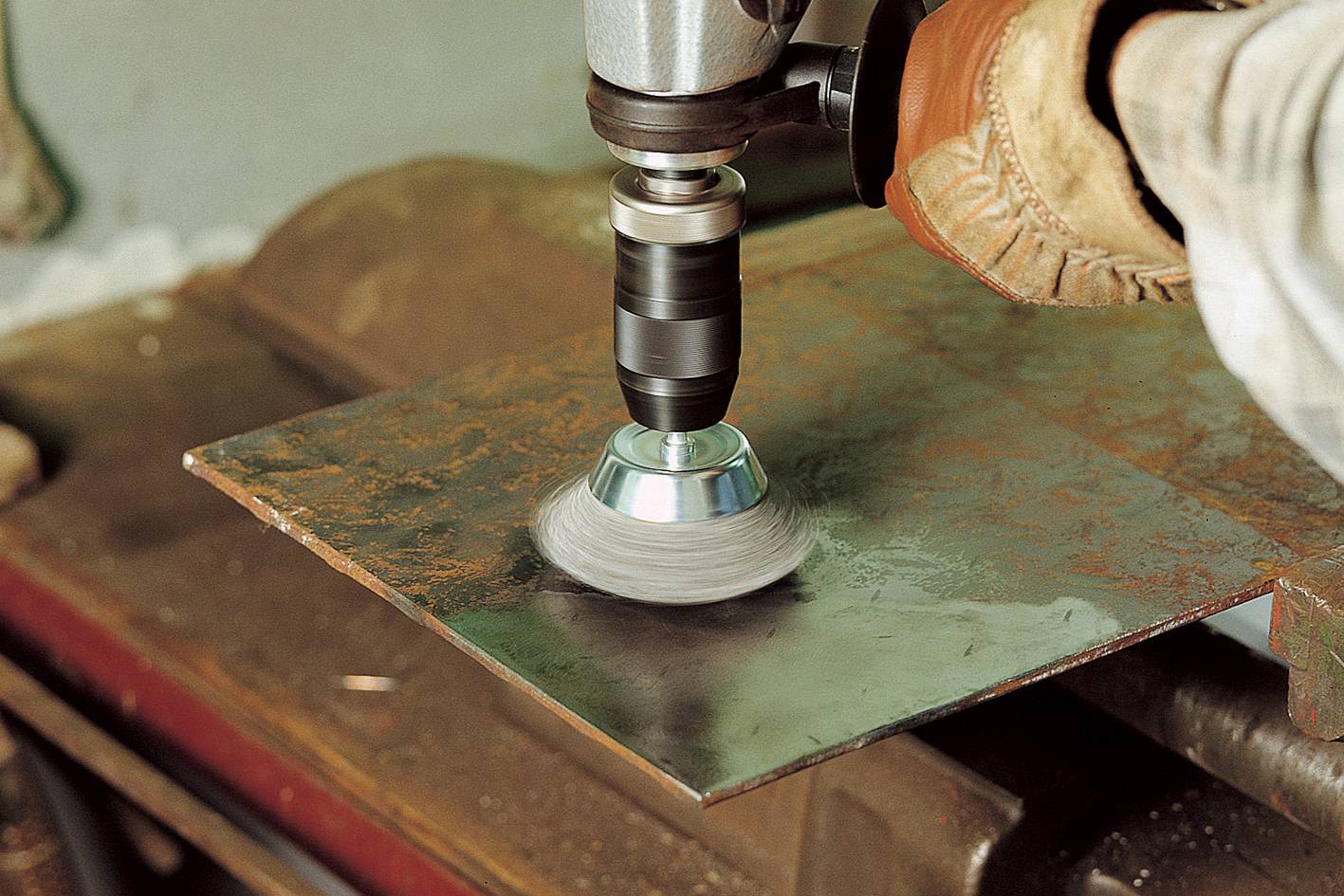 Osoba používa vŕtačku s drôtenou kefou, aby odstránila hrdzu z kovovej dosky. Má na sebe pracovné rukavice.