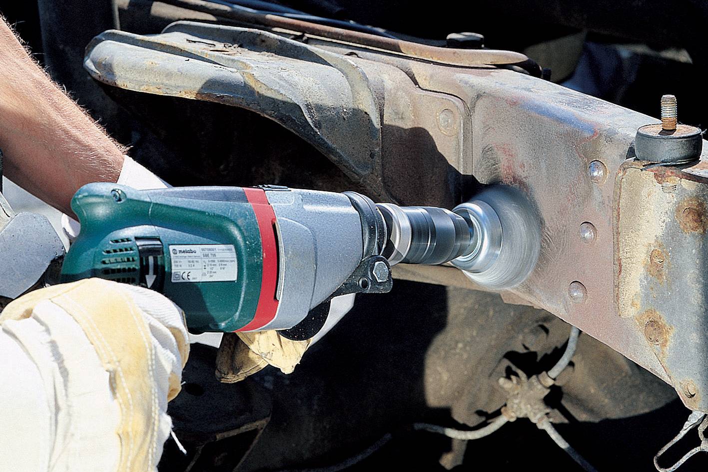 Osoba obrúsava hrdzu z kovového rámu elektrickým brúsnym strojom, pravdepodobne na vozidle.
