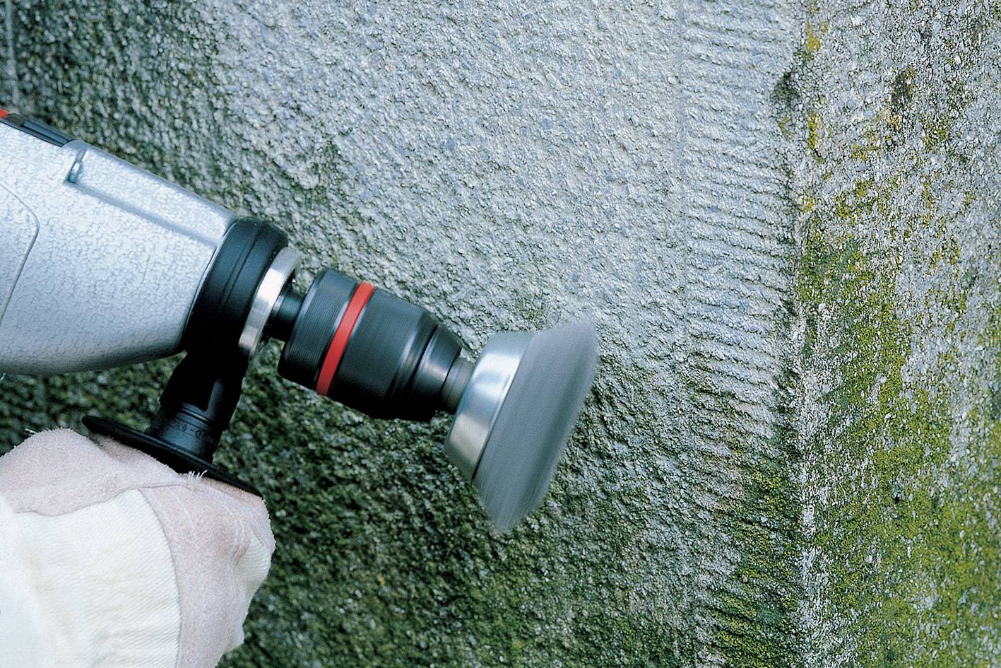 Jedna ruka drží stroj na brúsenie betónovej steny. Stena vykazuje známky machového porastu a opotrebenia.