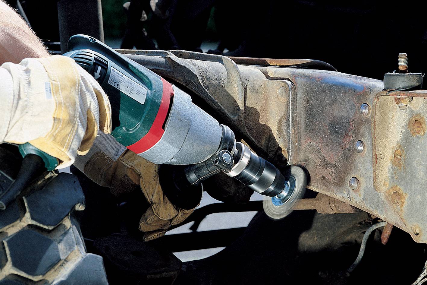 Osoba brúsi hrdzavý rám vozidla uhlovou brúskou. Má pritom pracovné rukavice.